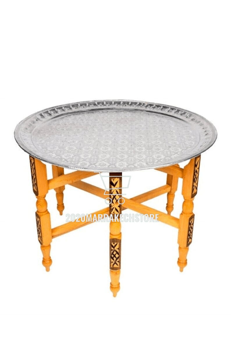 May include: A round silver tray table with a decorative pattern on the tray. The table has a wooden base with intricate carvings and four legs.