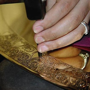 May include: A person using a hammer to create intricate designs on a gold metal surface. The surface is decorated with a floral pattern.