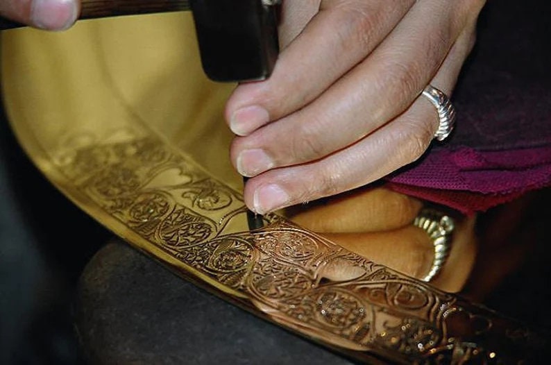 May include: A person using a hammer to create intricate designs on a gold metal surface. The surface is decorated with a floral pattern.