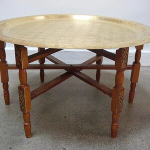 May include: A round brass tray table with a carved wooden base. The tray is hammered and has a shiny finish. The base has four legs with intricate carvings.
