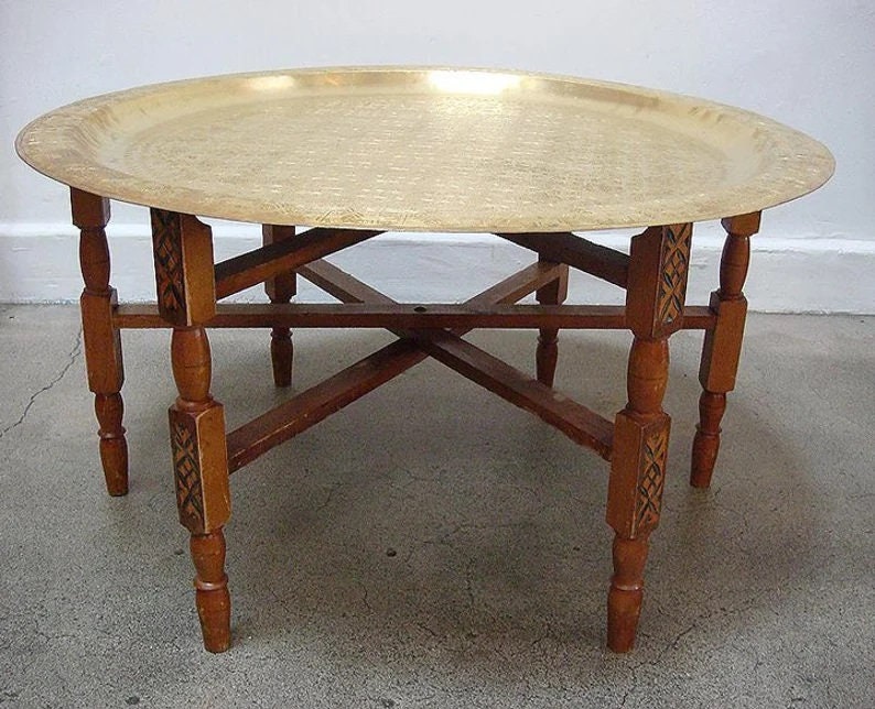 May include: A round brass tray table with a carved wooden base. The tray is hammered and has a shiny finish. The base has four legs with intricate carvings.