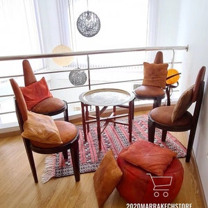 May include: A Moroccan-style seating area with four chairs, a round table with a silver tray, and a pouf. The chairs are made of wood and have a brown leather upholstery. The pouf is also made of brown leather. The table is made of wood and has a silver tray on top. The seating area is decorated with a rug with a geometric pattern. The room is decorated with a white wall and a window with a view of the outside.