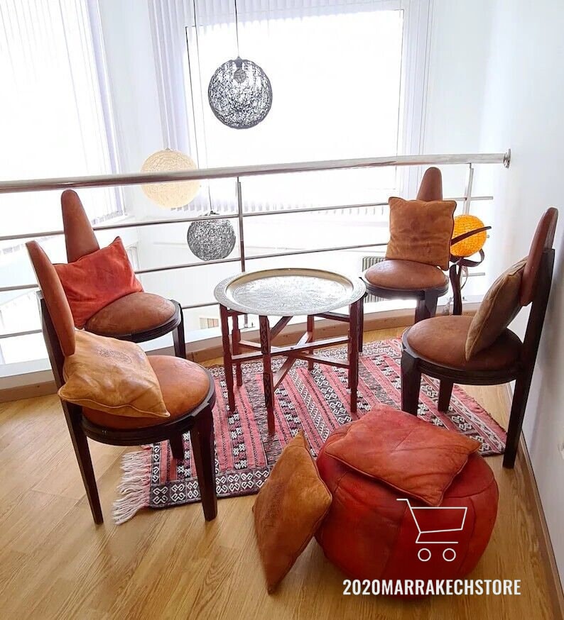 May include: A Moroccan-style seating area with four chairs, a round table with a silver tray, and a pouf. The chairs are made of wood and have a brown leather upholstery. The pouf is also made of brown leather. The table is made of wood and has a silver tray on top. The seating area is decorated with a rug with a geometric pattern. The room is decorated with a white wall and a window with a view of the outside.