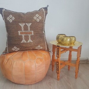 May include: A brown and white patterned pillow sits on top of a tan leather pouf. A small wooden table with a brass top holds three gold candle holders.