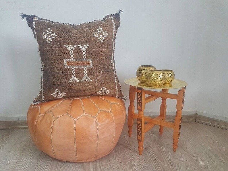 May include: A brown and white patterned pillow sits on top of a tan leather pouf. A small wooden table with a brass top holds three gold candle holders.