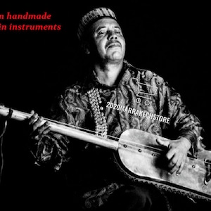 May include: A black and white image of a man playing a Moroccan instrument. The instrument is a stringed instrument with a long neck and a rounded body. The man is wearing a patterned shirt and a hat. Text in the image reads "African handmade Moroccan instruments" and "2020marrakechstore".