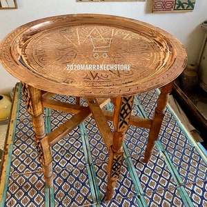 May include: A round copper tray with intricate engraved designs sits on a wooden stand with carved legs. The tray is on a tiled floor with a blue and yellow pattern.