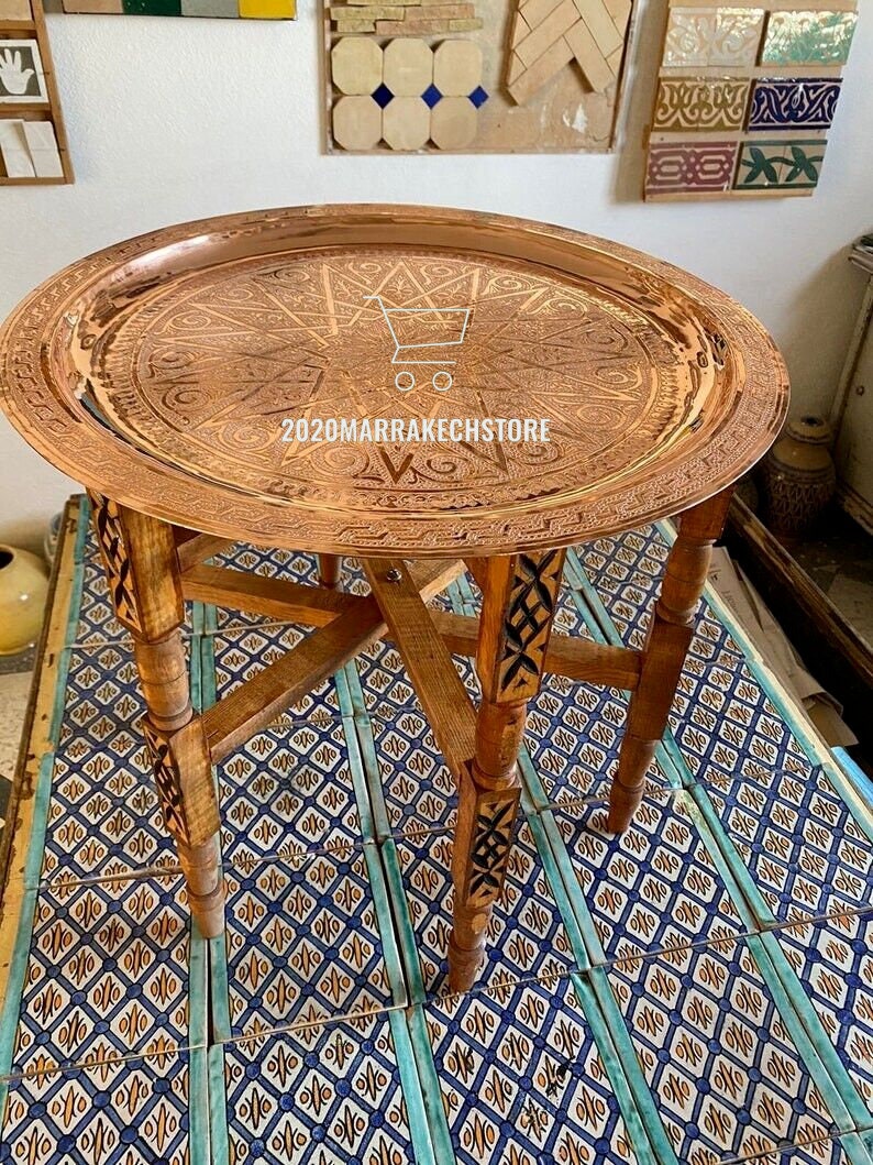 May include: A round copper tray with intricate engraved designs sits on a wooden stand with carved legs. The tray is on a tiled floor with a blue and yellow pattern.