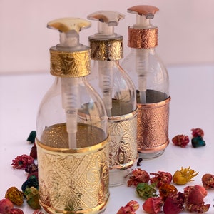 May include: Three clear glass bottles with decorative metal bands and pump dispensers. The metal bands are gold, silver, and copper-colored, with ornate embossed patterns. The bottles are surrounded by colorful dried flowers.