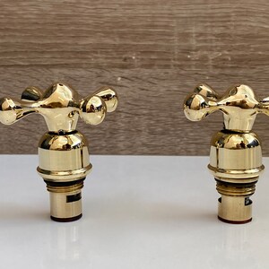 May include: Two gold-toned faucet handles with cross-shaped knobs. The handles are attached to a round base with a threaded stem.