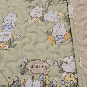 May include: A quilted, light green fabric featuring a repeating pattern of cartoon bunnies in a garden setting. The bunnies are depicted with white fur and wearing various outfits. The word "GARDEN" is on a sign. The fabric is bordered with a light green trim.