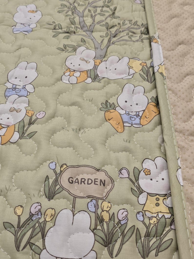 May include: A quilted, light green fabric featuring a repeating pattern of cartoon bunnies in a garden setting. The bunnies are depicted with white fur and wearing various outfits. The word "GARDEN" is on a sign. The fabric is bordered with a light green trim.