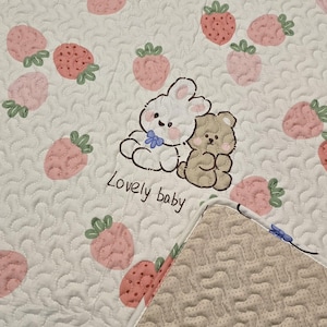 May include: White quilted baby blanket with a pattern of pink strawberries and the words "Lovely baby". The blanket features a cartoon rabbit and bear illustration. The reverse side has a beige, textured pattern.