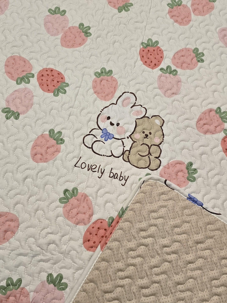 May include: White quilted baby blanket with a pattern of pink strawberries and the words "Lovely baby". The blanket features a cartoon rabbit and bear illustration. The reverse side has a beige, textured pattern.