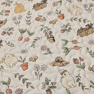 May include: A quilted fabric featuring a repeating pattern of bunnies, flowers, vegetables, and gardening tools. The design includes carrots, strawberries, mushrooms, and flower baskets in soft pastel colors. The background is a light cream color with a quilted texture.