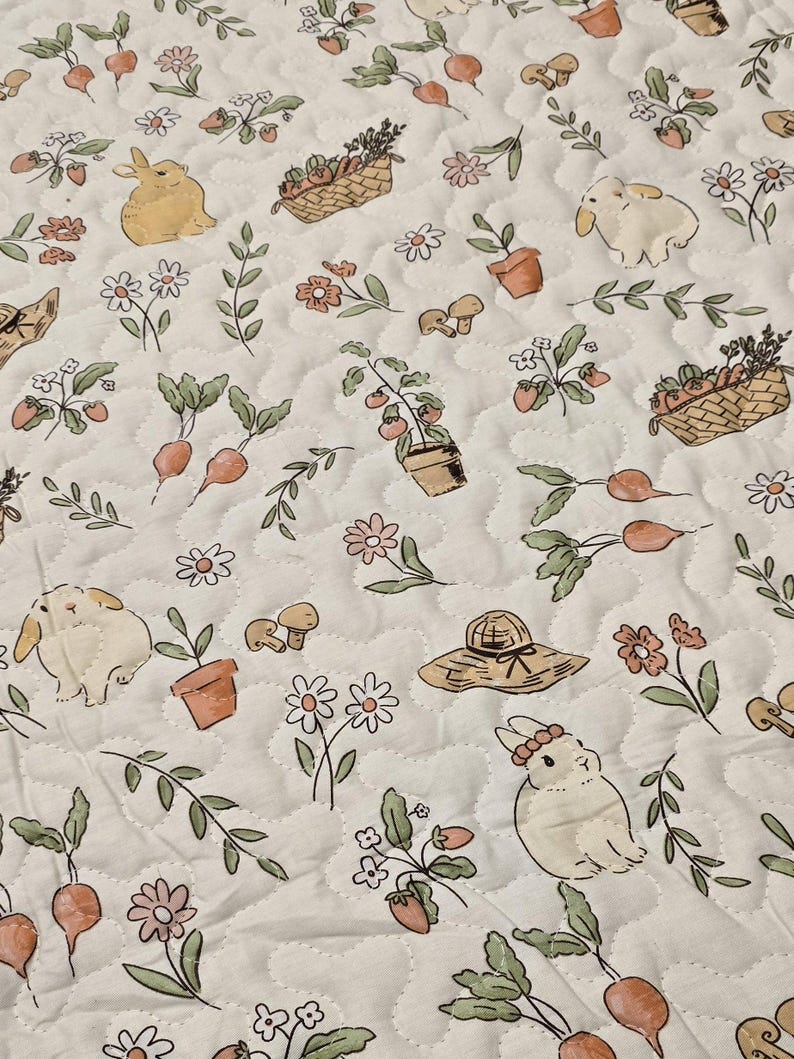 May include: A quilted fabric featuring a repeating pattern of bunnies, flowers, vegetables, and gardening tools. The design includes carrots, strawberries, mushrooms, and flower baskets in soft pastel colors. The background is a light cream color with a quilted texture.