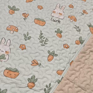 May include: A light green quilted fabric with a repeating pattern of cartoon bunnies, carrots, mushrooms, and flowers. The word "luck" is written near a bunny. The fabric has a wavy, stitched design.