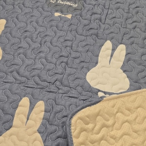 May include: A blue quilted blanket with a repeating pattern of white bunny silhouettes. The blanket has a beige border and the text "is turning" is visible. The blanket is made of a soft material and is suitable for a baby or child.