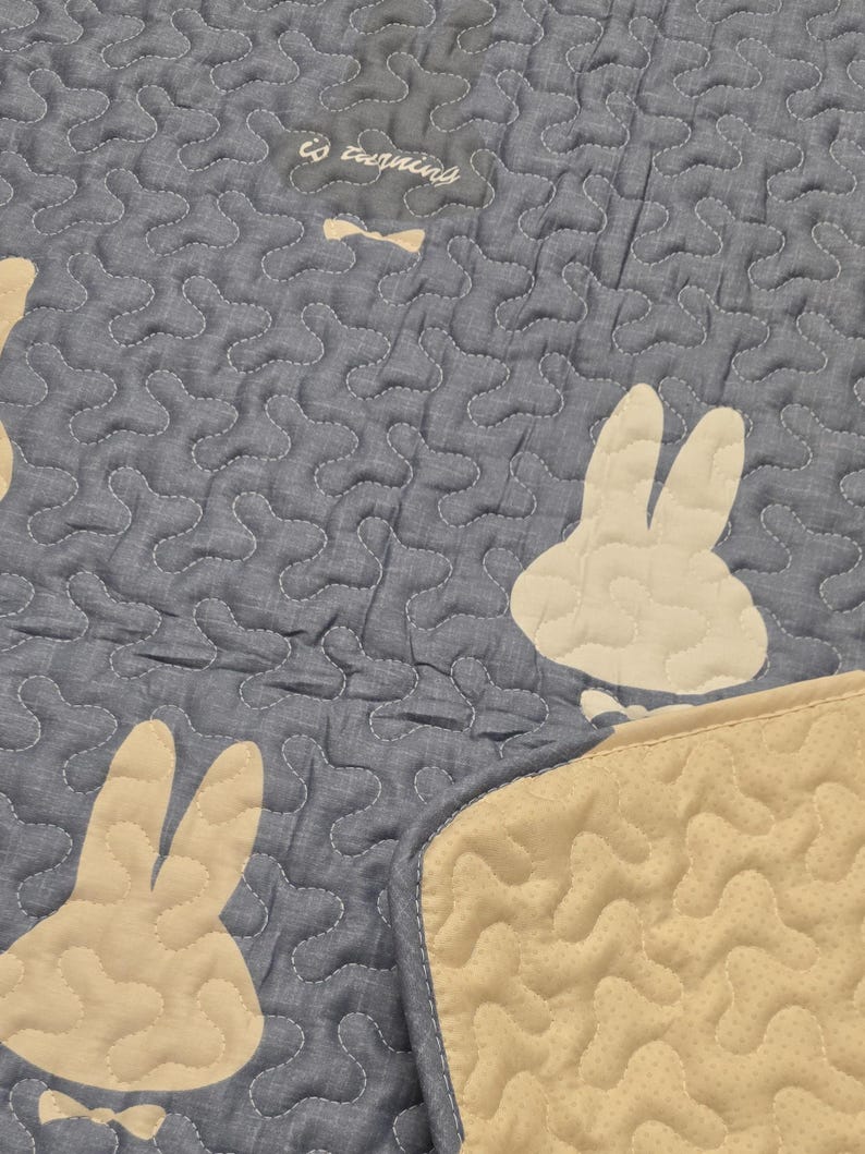 May include: A blue quilted blanket with a repeating pattern of white bunny silhouettes. The blanket has a beige border and the text "is turning" is visible. The blanket is made of a soft material and is suitable for a baby or child.