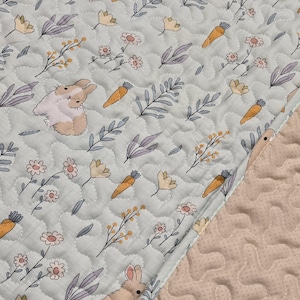 May include: A light blue quilted fabric with a repeating pattern of bunnies, carrots, flowers, and leaves. The bunnies are beige and white, the carrots are orange, and the flowers are yellow and pink. The edge of the fabric is a light beige color.