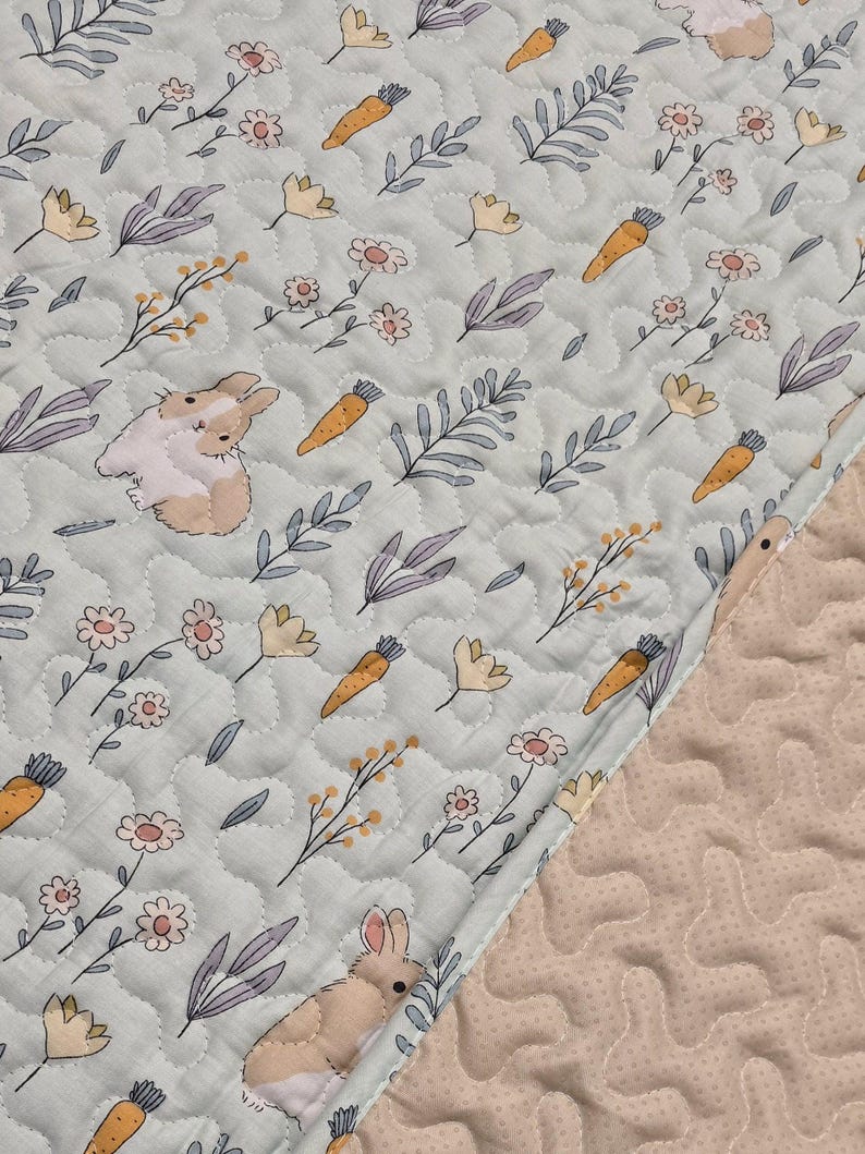 May include: A light blue quilted fabric with a repeating pattern of bunnies, carrots, flowers, and leaves. The bunnies are beige and white, the carrots are orange, and the flowers are yellow and pink. The edge of the fabric is a light beige color.