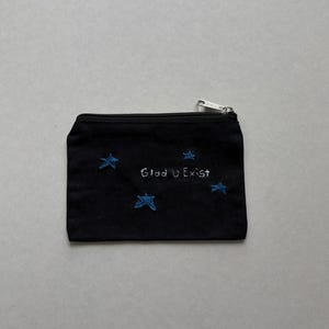 May include: Black zippered pouch with embroidered blue stars and the text "Glad U Exist". The small bag has a silver zipper and is likely made of a durable fabric. A simple and sweet accessory.