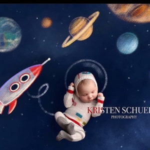 May include: A baby wearing a white astronaut costume with a red and white hat is sleeping in a white circle drawn on a dark blue background. The background also features a white rocket, a white ring around a yellow planet, and several other planets in various colours.