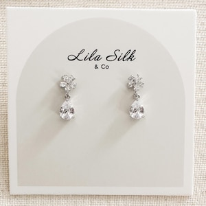 May include: A pair of silver teardrop earrings with a flower design. The earrings are on a white card with the text "Lila Silk & Co."