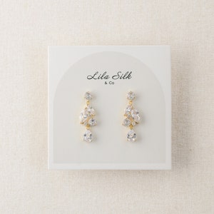 May include: A pair of gold dangle earrings with clear crystal stones. The earrings are displayed on a white card with the text "Lila Silk & Co."
