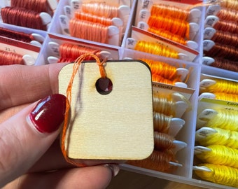 May include: A close-up of a wooden thread organizer with a variety of colored embroidery floss spools. The spools are labeled with names like "Peach", "Apricot", "Pumpkin", and "Lemon".