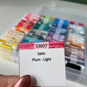 May include: A close-up of a small white card with the text "S3607 Satin Plum - Light" printed on it. The card is sitting on top of a clear plastic box filled with colorful spools of thread.