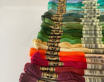 Puede incluir: Una colección de carretes de hilo de bordar DMC en varios colores, incluyendo rosa, morado, azul, verde, amarillo, naranja, rojo, marrón y gris. Cada carrete tiene una etiqueta dorada con el logotipo de DMC y el número del color.