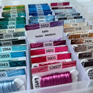May include: A close-up of a plastic storage box filled with spools of embroidery floss. The spools are labeled with numbers and colors. The floss is organized by color and type. The box is white and has a clear lid. The floss is a variety of colors, including pink, red, blue, green, and brown.