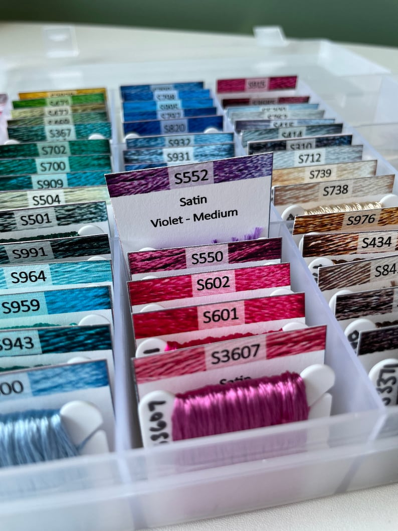 May include: A close-up of a plastic storage box filled with spools of embroidery floss. The spools are labeled with numbers and colors. The floss is organized by color and type. The box is white and has a clear lid. The floss is a variety of colors, including pink, red, blue, green, and brown.