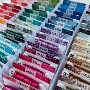 May include: A close-up of a plastic storage box filled with rows of colorful embroidery floss spools. Each spool is labeled with a number and the word "Satin".