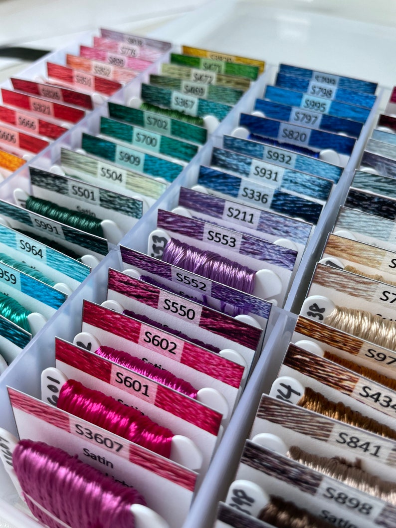 May include: A close-up of a plastic storage box filled with rows of colorful embroidery floss spools. Each spool is labeled with a number and the word "Satin".