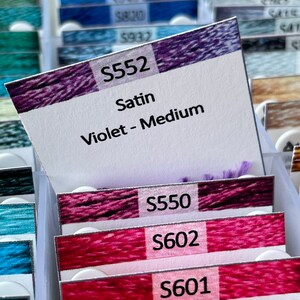 May include: A close-up of a collection of embroidery floss spools, with a label for spool number S552, Satin Violet - Medium.