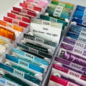 May include: Close-up of a white plastic storage box filled with spools of embroidery floss. The spools are organized by color and labeled with numbers and names. The floss is in shades of blue, green, purple, and pink. The labels include "Satin", "Blue Green - Very Light", and "S504".