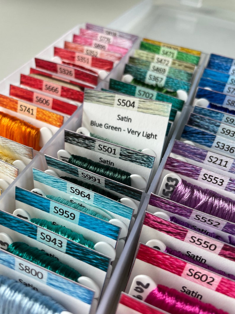 May include: Close-up of a white plastic storage box filled with spools of embroidery floss. The spools are organized by color and labeled with numbers and names. The floss is in shades of blue, green, purple, and pink. The labels include "Satin", "Blue Green - Very Light", and "S504".