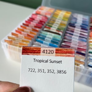 Op de afbeelding: Een witte kaart met de tekst "4120 Tropical Sunset 722, 351, 352, 3856" erop gedrukt. De kaart heeft een rood-oranje gestreepte achtergrond.