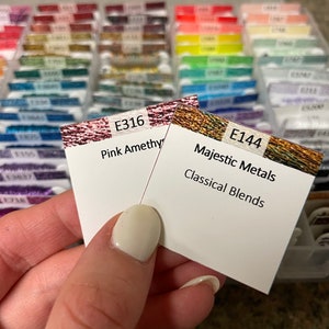 May include: Two DMC embroidery floss cards, one is pink amethyst with the number E316 and the other is gold with the number E144 and the text "Majestic Metals Classical Blends".