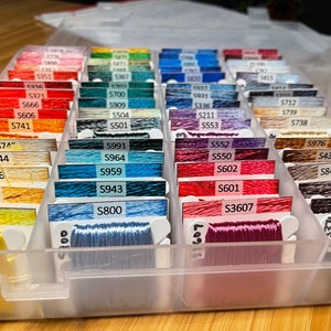 May include: A clear plastic storage box filled with rows of colorful embroidery floss spools. Each spool is labeled with a number, such as "S991", "S964", "S959", "S943", "S800", "S552", "S550", "S602", "S601", "S3607", "S976", "S434", "S841", "S898", "S3371", "S747", "S744", "S726", "S676", "S307", "S321", "S666", "S606", "S741", "S712", "S739", "S738", "C037", "S700", "0931", "S909", "S336", "S504", "S211", "S501", "S553", "5473", "5351", "5367", "5820", "5415".