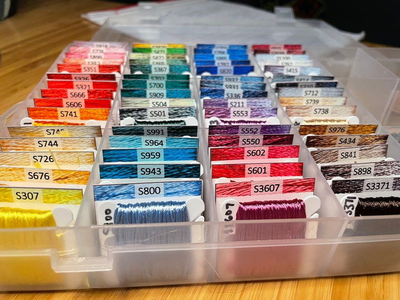 May include: A clear plastic storage box filled with rows of colorful embroidery floss spools. Each spool is labeled with a number, such as "S991", "S964", "S959", "S943", "S800", "S552", "S550", "S602", "S601", "S3607", "S976", "S434", "S841", "S898", "S3371", "S747", "S744", "S726", "S676", "S307", "S321", "S666", "S606", "S741", "S712", "S739", "S738", "C037", "S700", "0931", "S909", "S336", "S504", "S211", "S501", "S553", "5473", "5351", "5367", "5820", "5415".