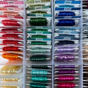 May include: A close-up of a plastic storage box filled with spools of embroidery floss. The spools are organized by color and labeled with numbers and names. The colors include red, orange, yellow, green, blue, purple, and pink.