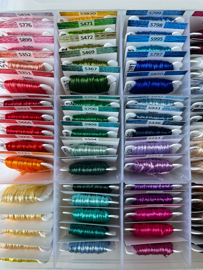 May include: A close-up of a plastic storage box filled with spools of embroidery floss. The spools are organized by color and labeled with numbers and names. The colors include red, orange, yellow, green, blue, purple, and pink.