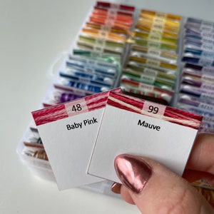 Puede incluir: Dos tarjetas de hilo de bordar con rayas rojas y blancas. La tarjeta de la izquierda está etiquetada como "48 Baby Pink" y la tarjeta de la derecha está etiquetada como "99 Mauve".