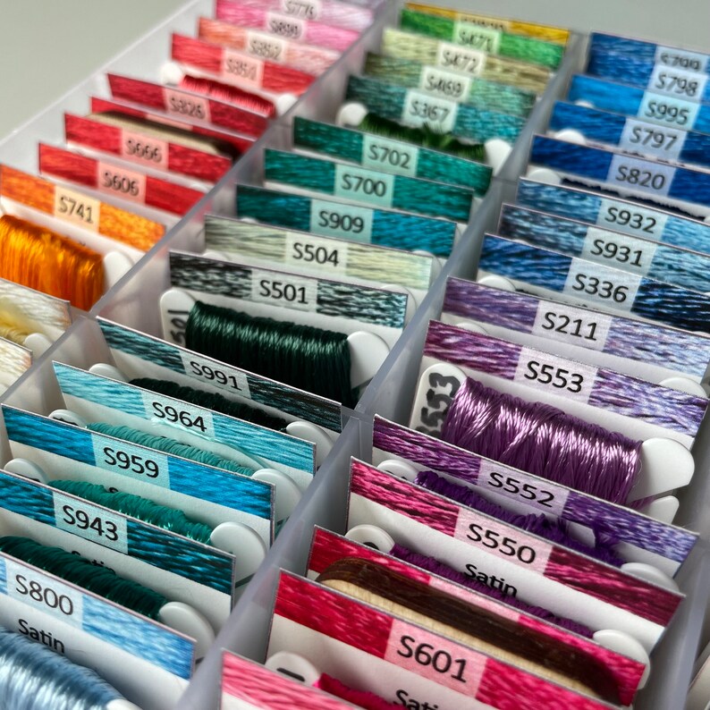 May include: A close-up of a plastic storage box filled with spools of embroidery floss. The spools are organized by color and labeled with numbers. The numbers on the spools include S741, S606, S501, S991, S964, S959, S943, S800, S553, S552, S550, S601, S702, S700, S909, S504, S469, S467, S471, S798, S797, S820, S932, S931, S336, S211.