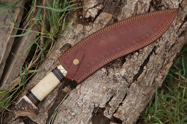 May include: A brown leather sheath with a decorative stitch pattern and a brass button. The sheath is wrapped around a knife with a white bone handle and a dark wood accent.