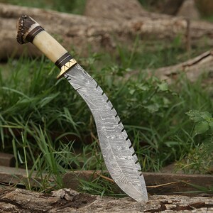 May include: A curved, silver-colored knife with a patterned blade and a brown and white handle. The handle is made of antler and has a gold-colored metal accent. The knife is resting on a piece of wood.