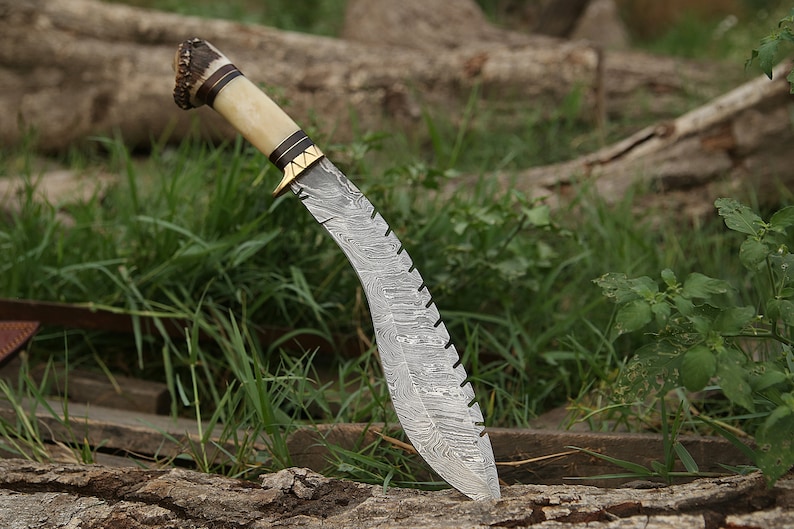 May include: A curved, silver-colored knife with a patterned blade and a brown and white handle. The handle is made of antler and has a gold-colored metal accent. The knife is resting on a piece of wood.